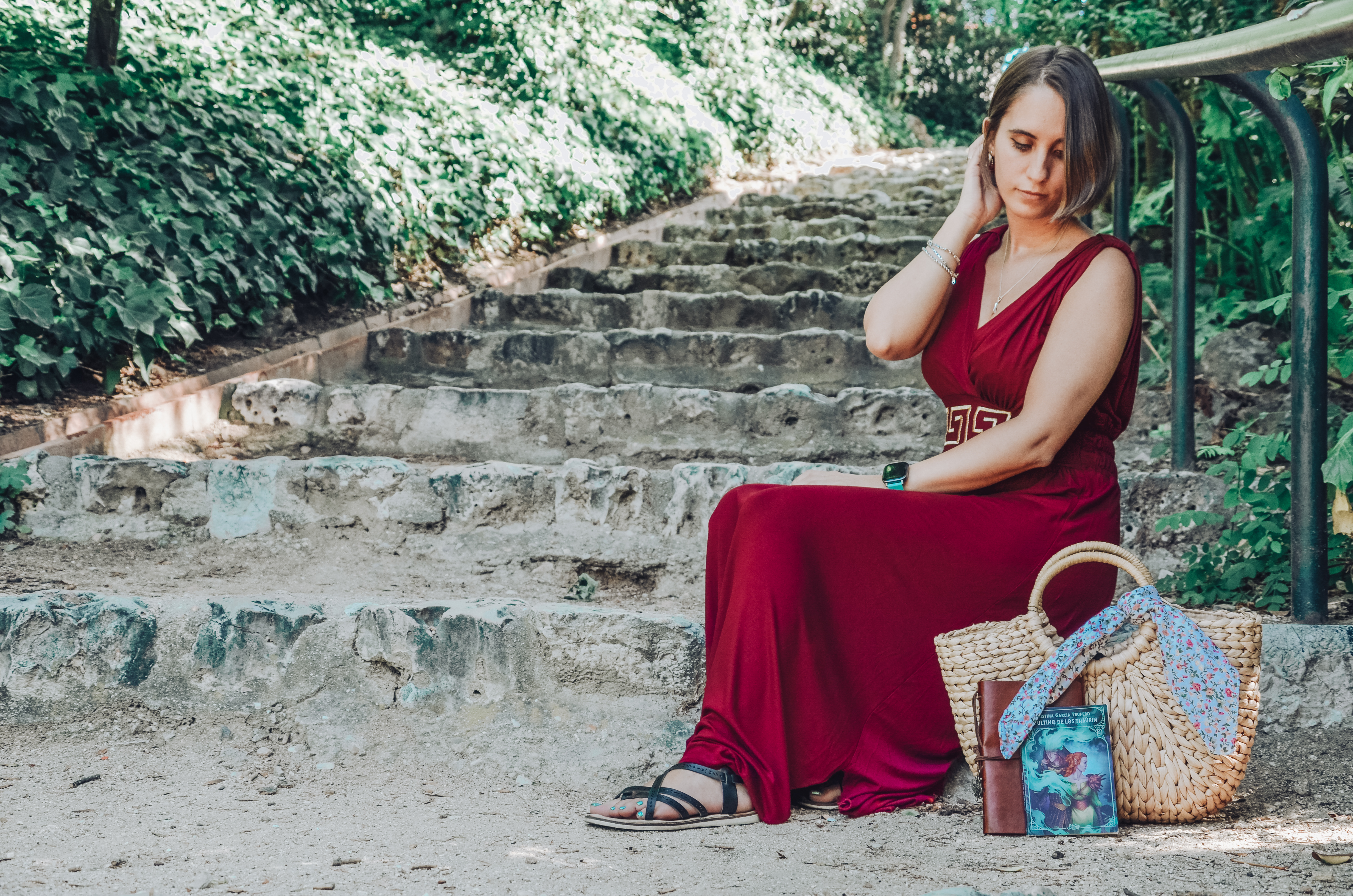 Fotografía en la que estoy sentada en unas escaleras con un vestido de corte griego color rojo oscuro. Tengo una mano en el pelo, para sujetarmelo y estoy mirando hacia abajo, hacia el bolso de mimbre que tengo delante y que tiene una libreta y la novelette apoyadas sobre él